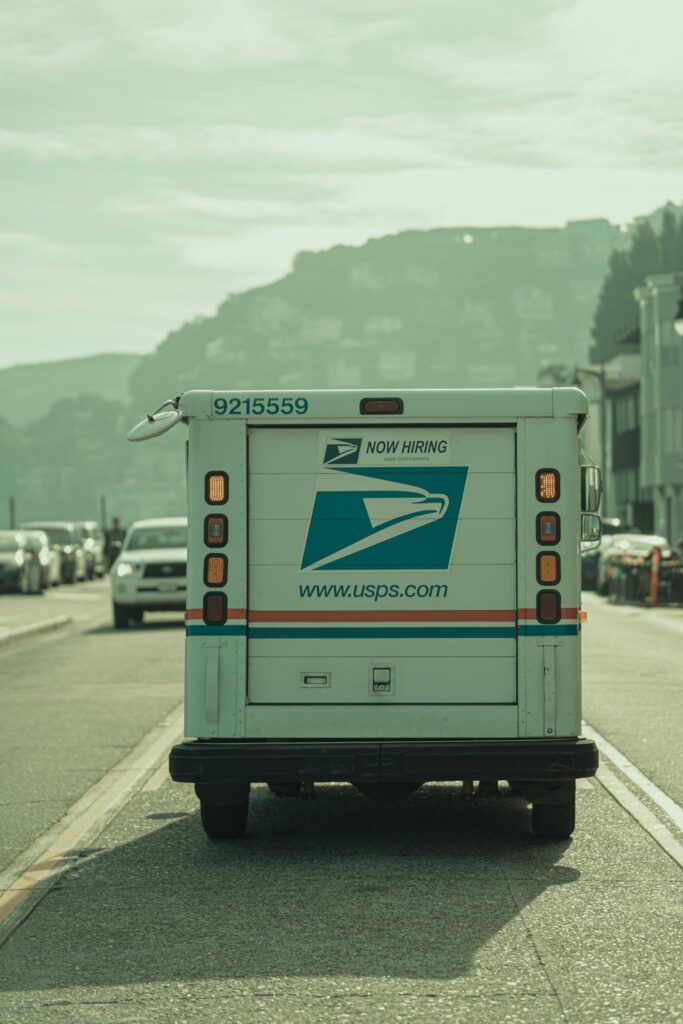 A USPS delivery truck driving down a scenic road in Sausalito, California, featuring hillside views.